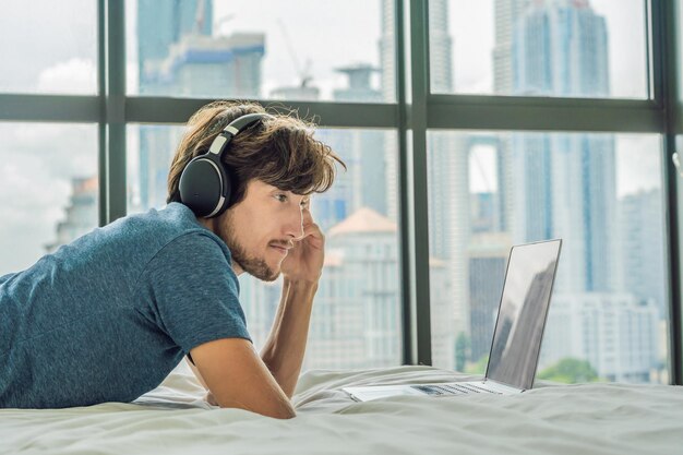 Young man is working on a laptop in his bed on a background of a panoramic window overlooking the skyscrapers. Uses wireless headphones. Freelancer, remote work, work from home