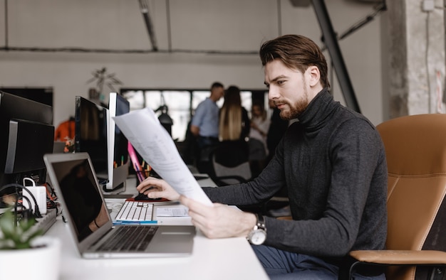 Young man is working at the laptop and making notes in the document sitting at desk in the modern office.