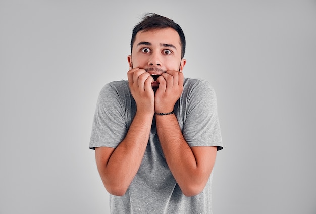 Young man isolated on grey background dressed in casual clothes, covering mouth with hands and round eyes experiencing deep astonishment and fear