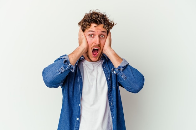 Young man isolated on white wall covering ears with hands trying not to hear too loud sound