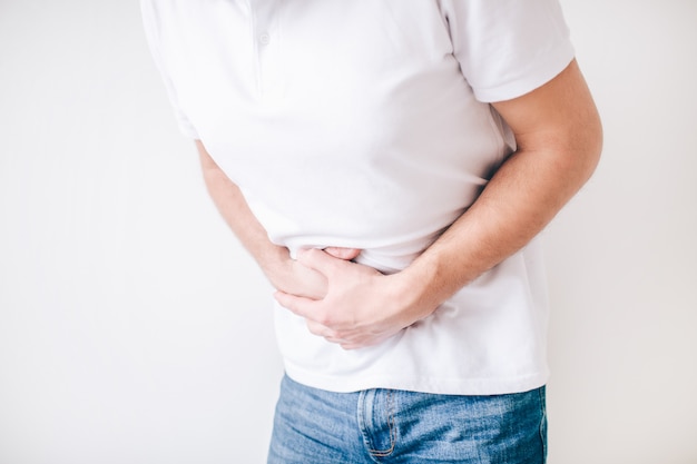 Young man isolated over white wall. Cut view of guy holding hands over stomach because of diarrhea. Painful and aching.