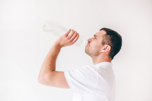 Young man isolated over white wall. Side view of guy drinking water from bottle.