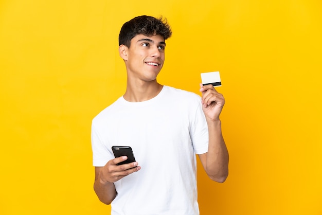 Young man over isolated yellow buying with the mobile with a credit card while thinking