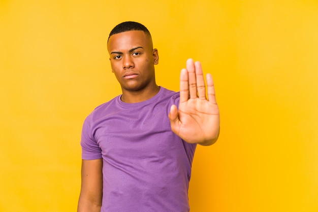 Young man isolated on yellow wall standing with outstretched hand showing stop sign, preventing you