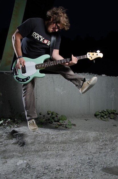 Photo young man jumping and playing bass guitar at night