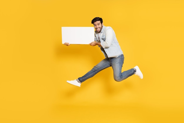 Young man jumping and showing blank white billboard isolated on yellow background