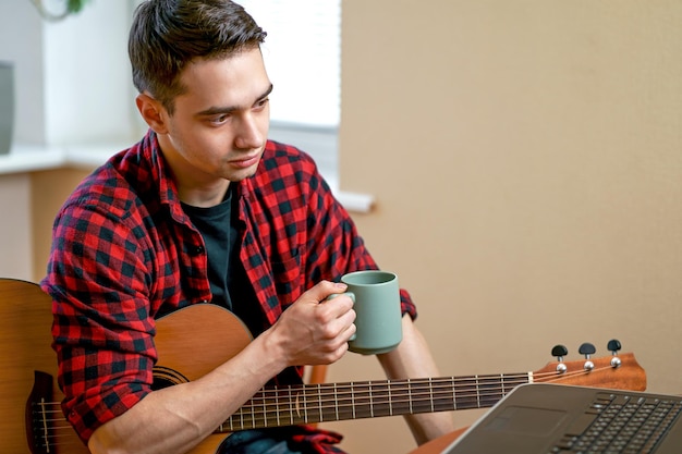 Young man learns to play the guitar takes lessons using the Internet sits near a coffee table with a cup of hot tea Workplace near the window Work at home