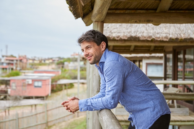 Young man in a light blue shirt leaning on the wooden railing of the terrace of his cabin while smiling.