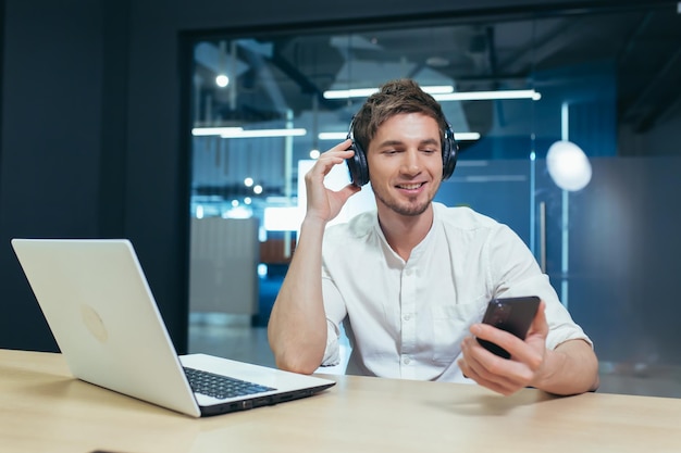 A young man listens to podcasts in headphones holds a mobile phone in his hands talks on video communication Sitting at work in the office with a laptop