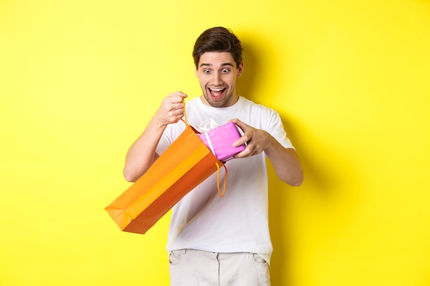 Young man looking surprised as take out gift from shopping bag, standing over yellow wall