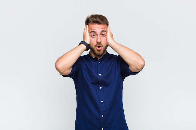 Young man looking unpleasantly shocked, scared or worried, mouth wide open and covering both ears with hands