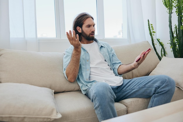 A young man looks angrily and sadly at the phone screen at home sitting on the couch Bad news and bad luck at work