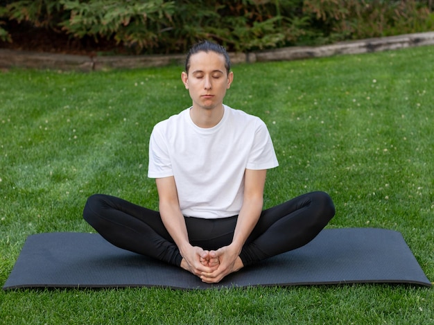 Young man in lotus position while practising yoga with his eyes closed on a black mat in the garden