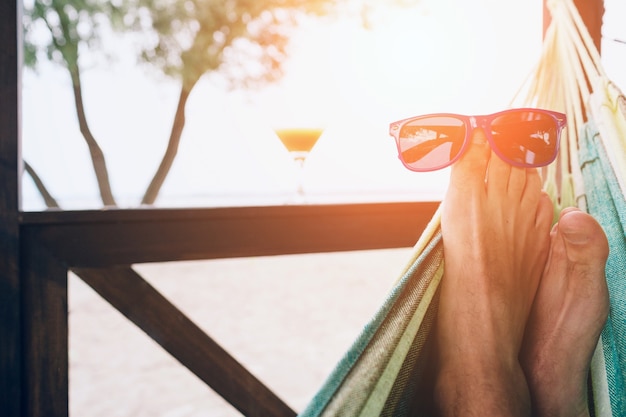 Young man lying in a hammock at a sunny beach by ocean. Sunglasses on the feet