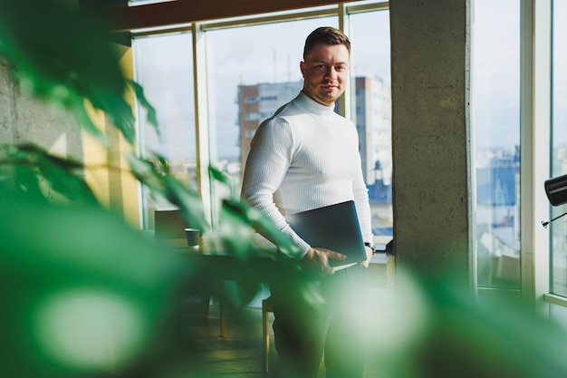 A young man in a modern workspace stands near a large window A man in casual clothes with a laptop in his hands Remote work Nice young manager