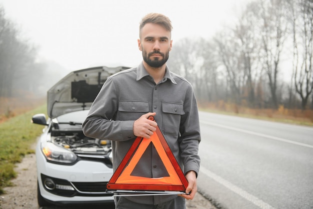 A young man near a broken car with an open hood on the roadside