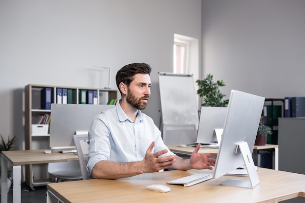 A young man an office worker sitting at a desk in the office at the computer talking on a video call to a computer camera discussing work with partners customers