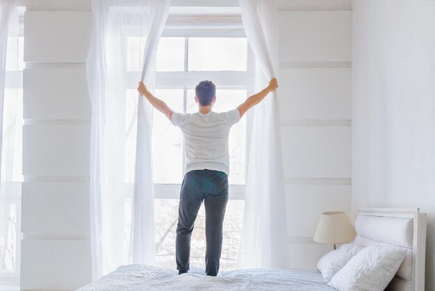 Young man opening window curtains in white bedroom in the morning