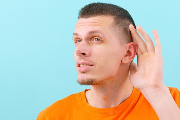 A young man in an orange shirt holding his hand near his ear and listening carefully over the blue background Silence Whispering Holding Carefully Casual Hearing Male Audience Sensing