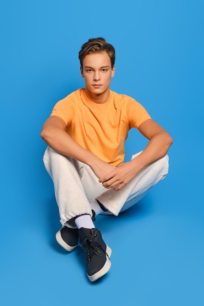 Young man in orange undershirt and white sweatpants sits on studio floor over blue background