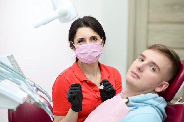 Young man patient is sitting at doctor dentist chair at dental office woman doctor in medical mask