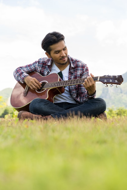 Young man plays guitar while sitting on green grass against nature and mountain landscape 