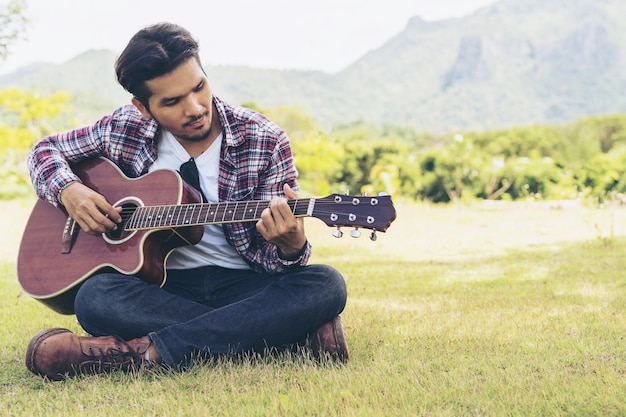 Young man plays guitar while sitting on green grass against nature and mountain landscape