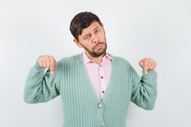 Young man pointing down in shirt, cardigan and looking dismal. front view.