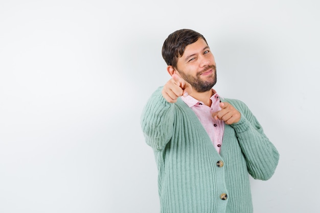 Young man pointing at front in shirt, cardigan and looking cheery , front view.