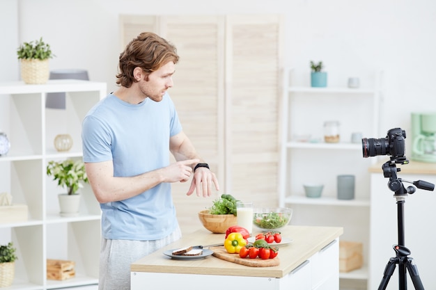 Young man pointing at his watch and telling about the time of healthy eating to the camera for his followers