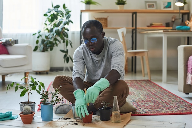 Young man in protective gloves sitting on the floor and planting plants into the pots in the room