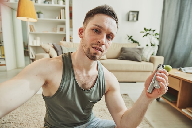 Young man recording himself while sitting on the floor in the center of living-room and doing exercise for hands and arm muscles