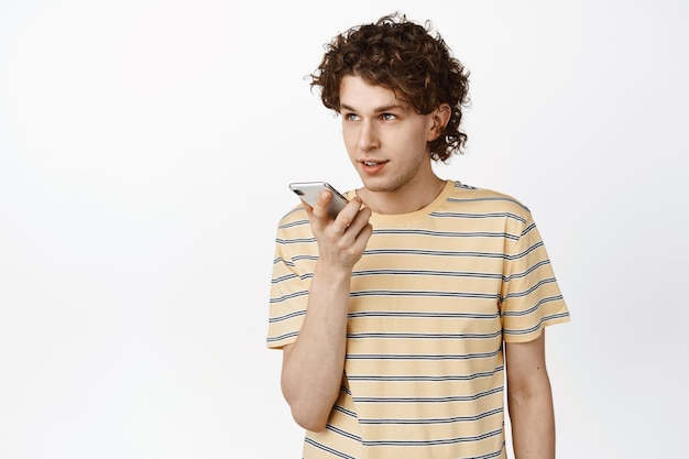 Young man recording voice message on mobile phone translating his speech with smartphone app standing over white background