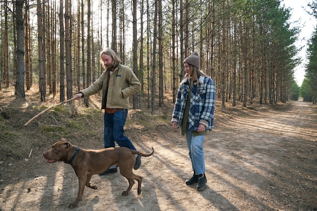 Young man showing stick to his dog and playing with him during walk in the park together with his girlfriend