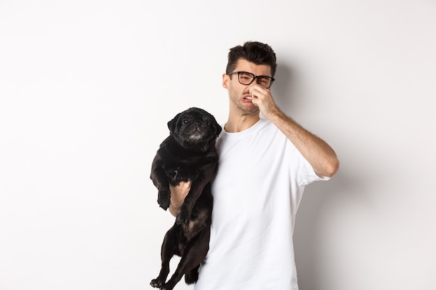 Young man shut nose as holding pug, disgusted with bad smell fart of animal, standing over white