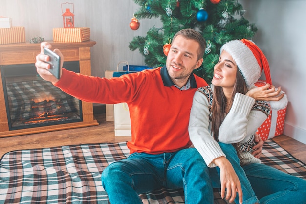 Young man sit on blanket with woman and takes selfie. He embrace her. She poses and smiles. 