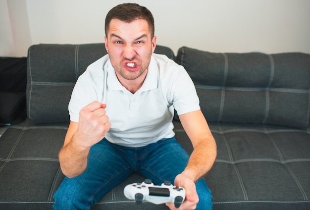 Young man sit in room during quarantine. Guy playing intense conputer game alone. Showing different rude emotions on his face.