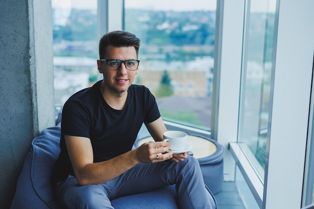 A young man sits on a bean bag chair near a large window in the office and drinks coffee during his lunch break A man in a black tshirt and trousers rests during lunch