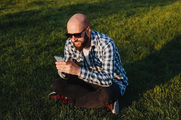 A young man sits on the green grass in the park and uses social networks using a mobile phone