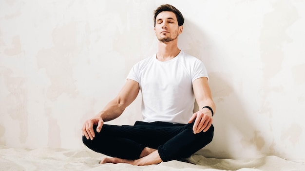 Young man sits in a meditative pose on sand against a wall background