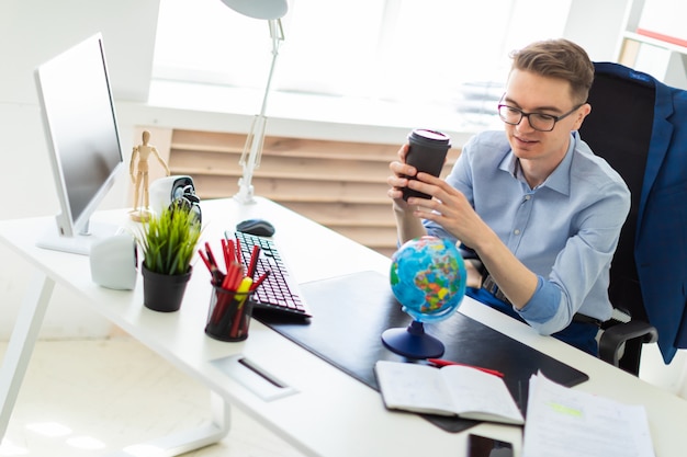 Photo a young man sits in the office at a computer desk and holds a glass of coffee in his hand. a young man faces a globe.