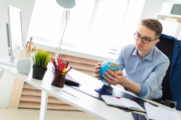 Photo a young man sits in the office at a computer desk and holds a globe in his hands.