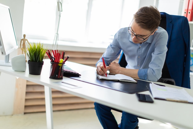 A young man sits in the office at a computer desk and writes in a notebook.