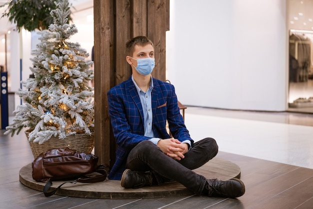 Young man sits in a shopping center in a medical mask with phone in his hand by the christmas tree