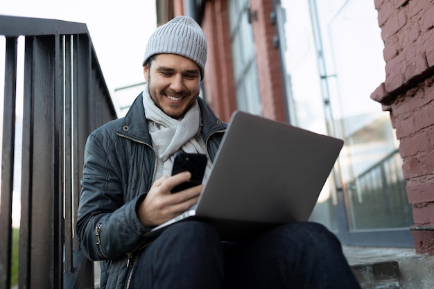 A young man sits on the steps with a laptop and looks at the phone with a smile