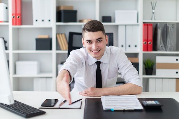 A young man sits at a table in the office and stretches his hand forward.