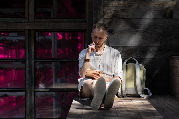 Young man sits on wooden stairs outdoors writing notes in paper pad in rays of sunlight