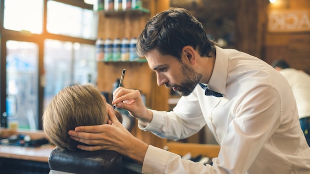 Young man sitting in a barbershop while barber working with razor