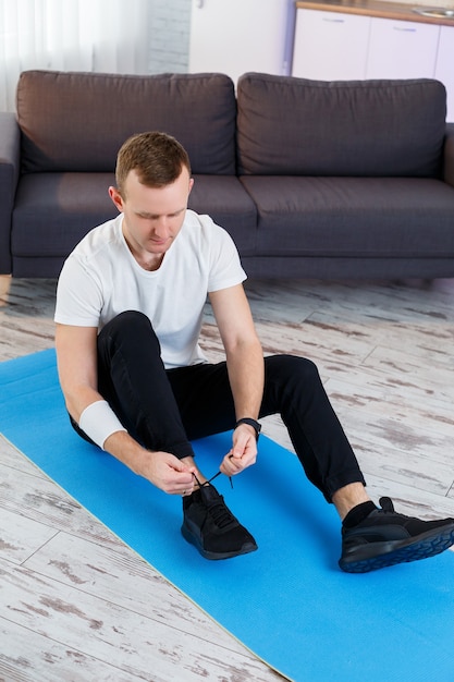 Young man sitting on carpet in his home and tying laces on sneakers after doing sports