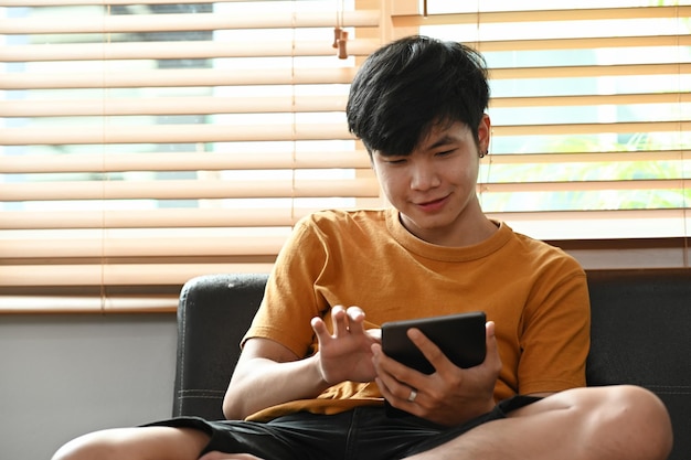 Young man sitting on comfortable couch and surfing internet with digital tablet